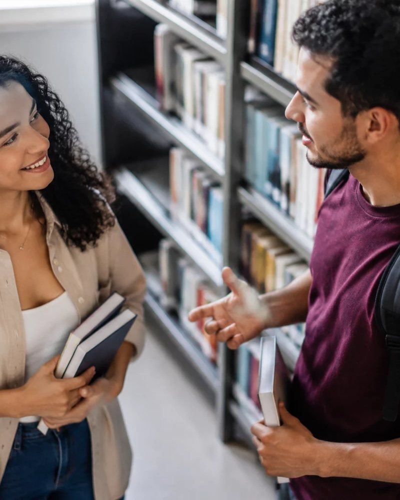 Duas participantes conversando de pé entre as prateleiras de uma biblioteca